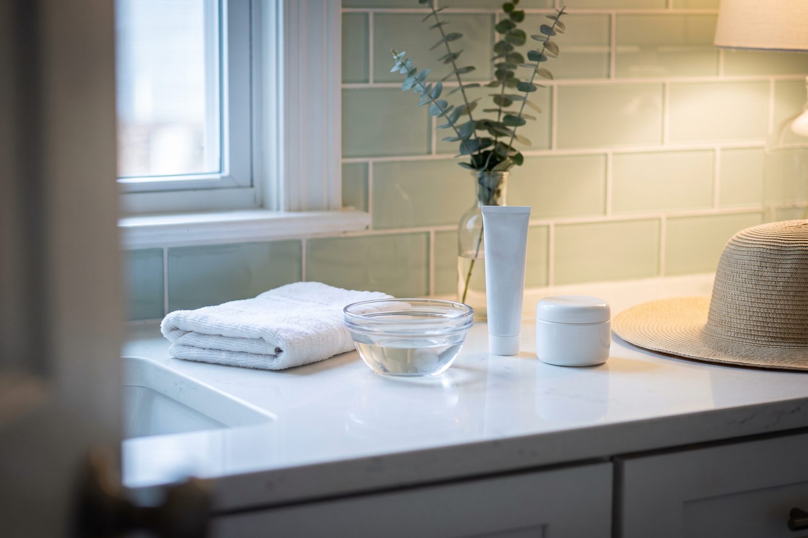 AI-generated illustration of a bathroom counter with a folded washcloth, a bowl of cool water, a plain jar of moisturizer and a straw hat