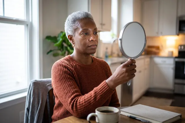 AI-generated illustration of a woman in her 50s at a kitchen table studying her cheek and jawline in a hand mirror