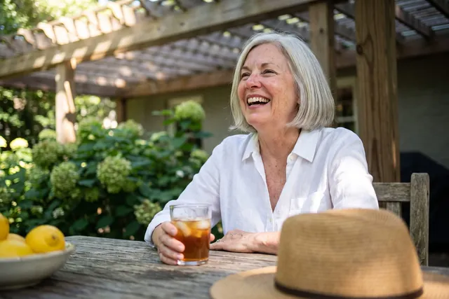 AI-generated illustration of a woman in her 60s with a silver bob laughing at a shaded patio table on a sunny morning