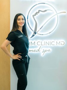 A professional photo of a female medical staff member in dark scrubs standing confidently in front of a clinic logo